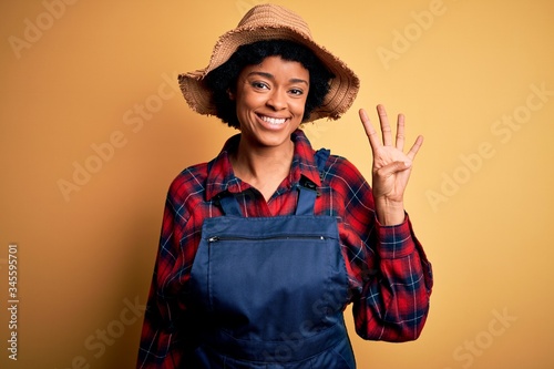 Young African American afro farmer woman with curly hair wearing apron and hat showing and pointing up with fingers number four while smiling confident and happy.