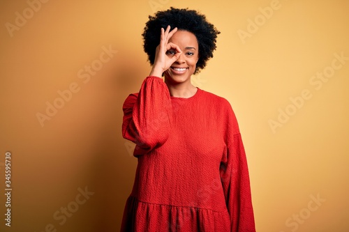 Young beautiful African American afro woman with curly hair wearing casual sweater doing ok gesture with hand smiling, eye looking through fingers with happy face.
