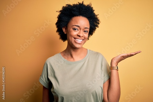 Young beautiful African American afro woman with curly hair wearing casual t-shirt smiling cheerful presenting and pointing with palm of hand looking at the camera.