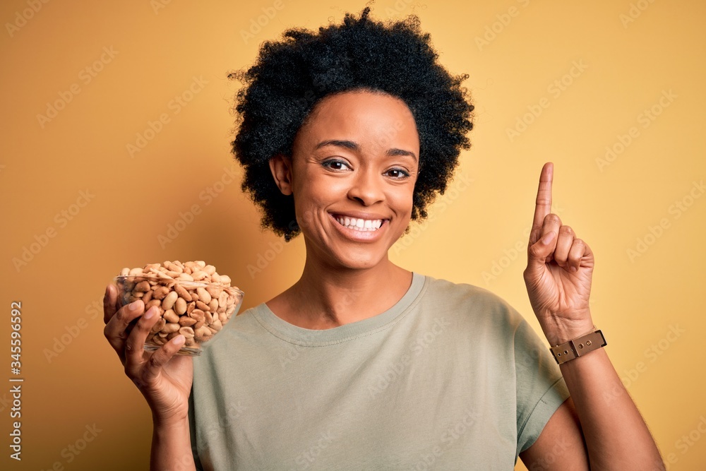 Young African American afro woman with curly hair holding bowl with salty peanuts surprised with an idea or question pointing finger with happy face, number one Young African American afro woman with curly hair holding bowl with salty peanuts surprised with an idea or question pointing finger with happy face, number one