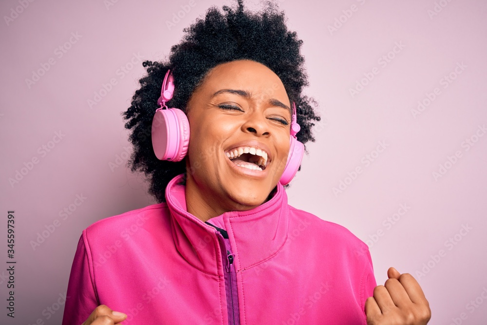 Young African American afro woman with curly hair listening to music using pink headphones very happy and excited doing winner gesture with arms raised, smiling and screaming for success. Celebration
