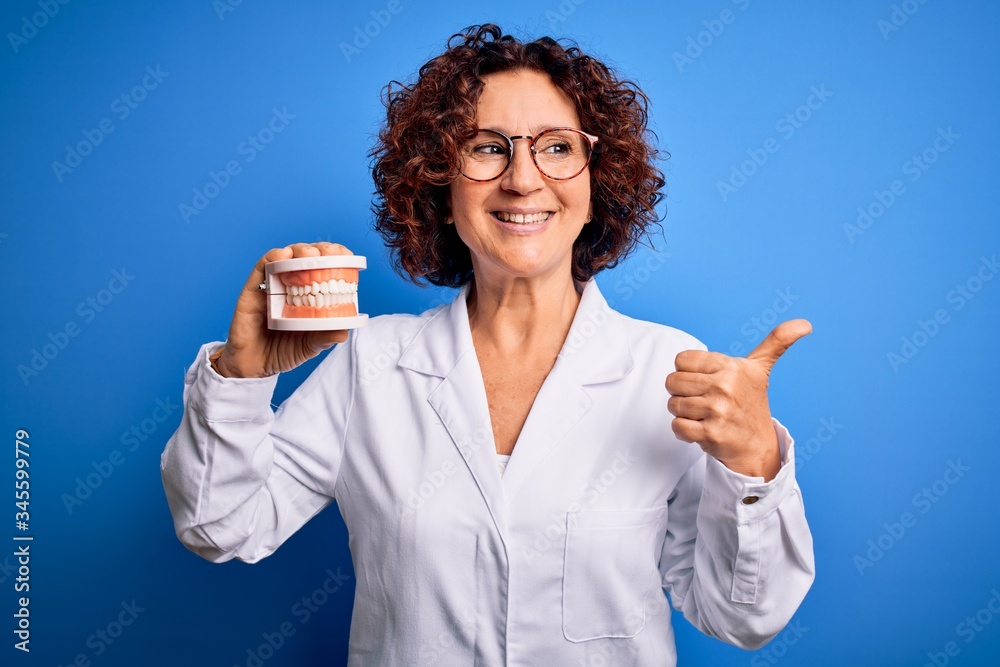 Middle age dentist woman wearing coat holding plastic denture teeth over blue background pointing and showing with thumb up to the side with happy face smiling Middle age dentist woman wearing coat holding plastic denture teeth over blue background pointing and showing with thumb up to the side with happy face smiling