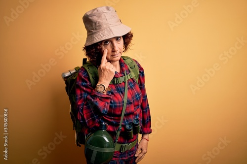 Middle age curly hair hiker woman hiking wearing backpack and water canteen using binoculars Pointing to the eye watching you gesture, suspicious expression