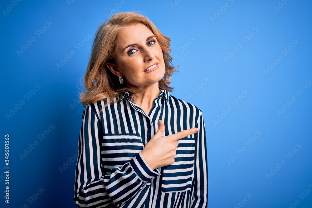 Middle age beautiful blonde woman wearing casual striped shirt standing over blue background cheerful with a smile of face pointing with hand and finger up to the side.