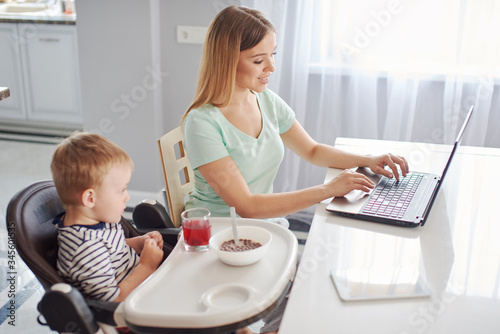 Young woman with child studying on laptop, making notes while feeding baby