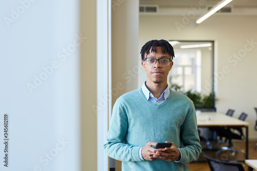 Horizontal waist up shot of handsome young Black man wearing shirt and blue sweater standing in office holding smartphone, looking at camera