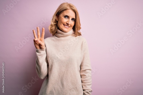 Valokuva Middle age beautiful blonde woman wearing casual turtleneck sweater over pink background showing and pointing up with fingers number three while smiling confident and happy