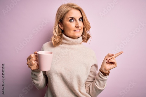 Middle age beautiful blonde woman drinking pink mug of coffee over isolated background very happy pointing with hand and finger to the side