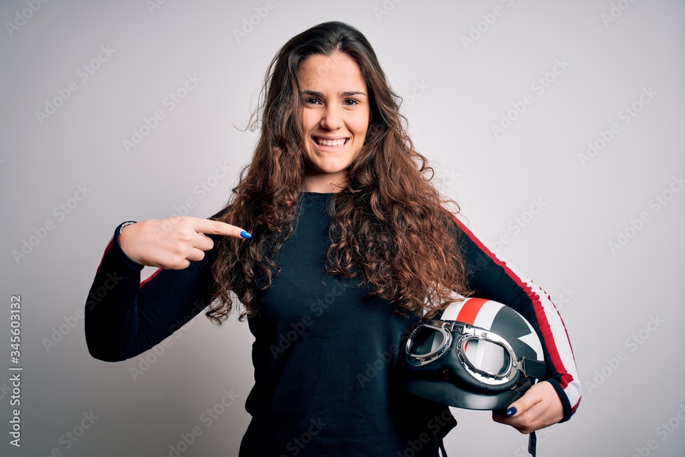 Obraz premium Beautiful motorcyclist woman with curly hair holding moto helmet over white background with surprise face pointing finger to himself