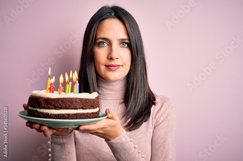 Young woman with blue eyes holding birthday cake with candles over pink background with a confident expression on smart face thinking serious