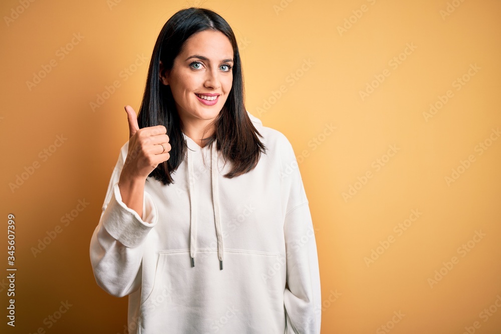 Young brunette sportswoman with blue eyes wearing training sweatshirt over yellow background doing happy thumbs up gesture with hand. Approving expression looking at the camera showing success. Young brunette sportswoman with blue eyes wearing training sweatshirt over yellow background doing happy thumbs up gesture with hand. Approving expression looking at the camera showing success.