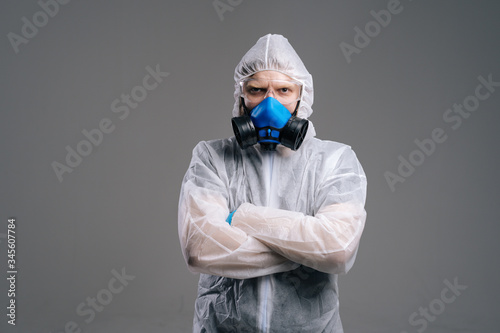 Serious epidemiologist medical worker in protective coveralls, glasses and respirator holding hands crossed on chest. Concept of Coronavirus COVID-19 Pandemic. Studio shot on isolated dark background.