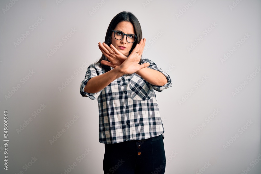Young brunette woman with blue eyes wearing casual shirt and glasses over white background Rejection expression crossing arms doing negative sign, angry face Young brunette woman with blue eyes wearing casual shirt and glasses over white background Rejection expression crossing arms doing negative sign, angry face