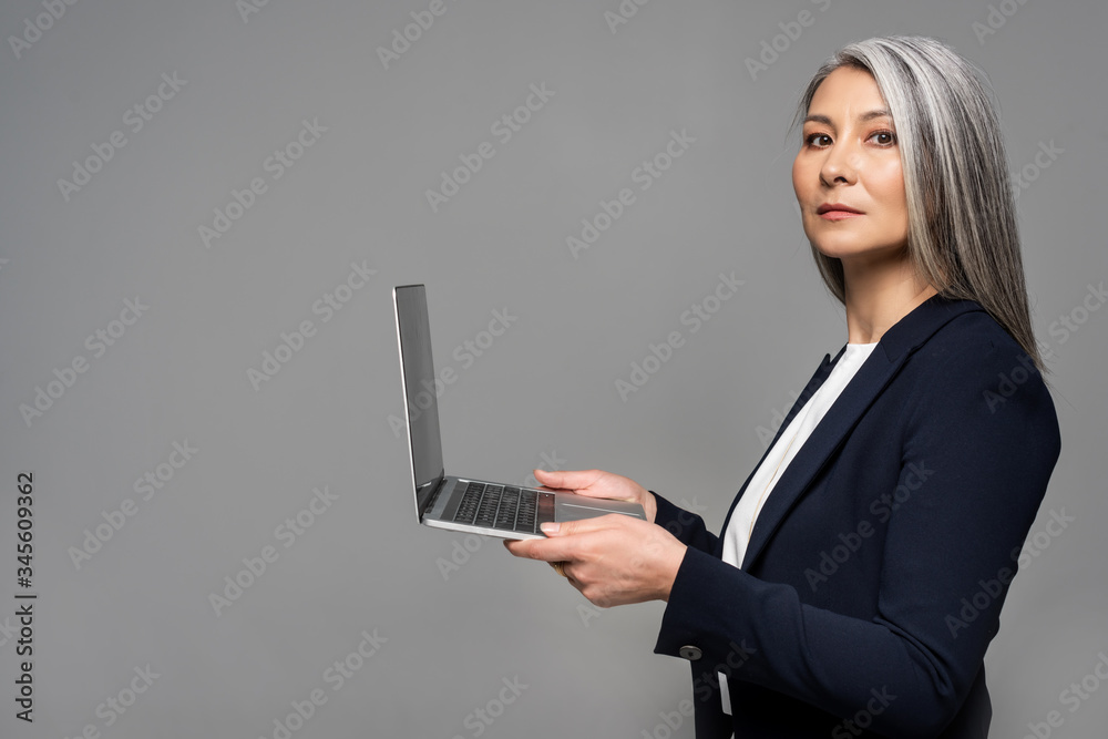 attractive serious asian businesswoman with grey hair using laptop isolated on grey attractive serious asian businesswoman with grey hair using laptop isolated on grey