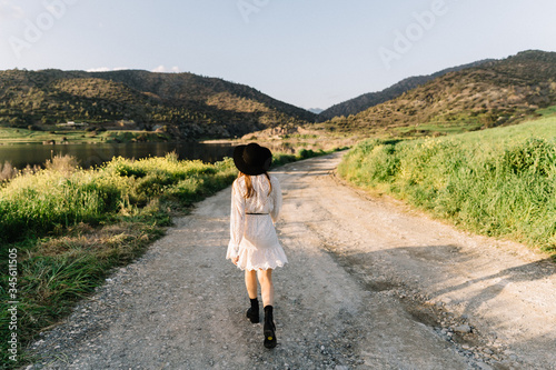 Young adorable girl in white dress and black hat, in spring, posing for camera. female portrait outdoor in mountain lake. Green fresh grass. Young unusual person enjoying nature