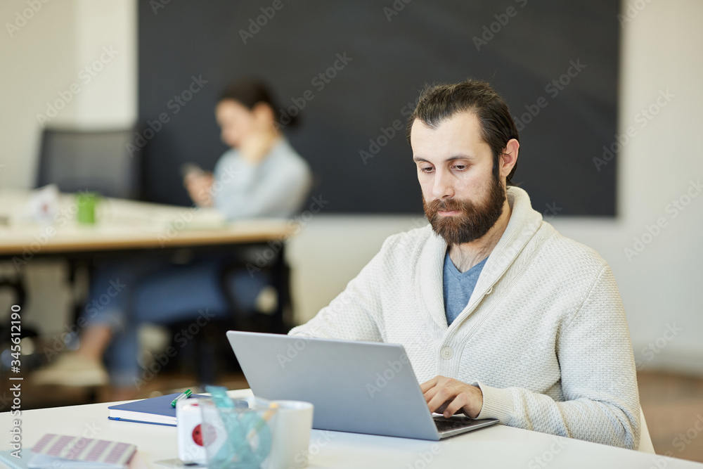Handsome young man with beard on face sitting at office table working on his laptop with serious facial expression