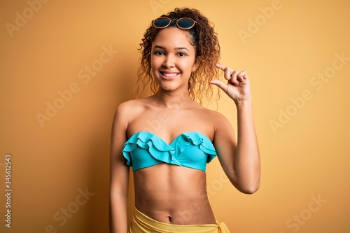 Obraz na plátne Young beautiful american woman on vacation wearing bikini over isolated yellow background smiling and confident gesturing with hand doing small size sign with fingers looking and the camera