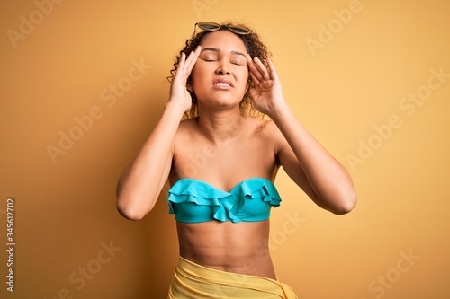 Young beautiful american woman on vacation wearing bikini over isolated yellow background with hand on headache because stress. Suffering migraine.