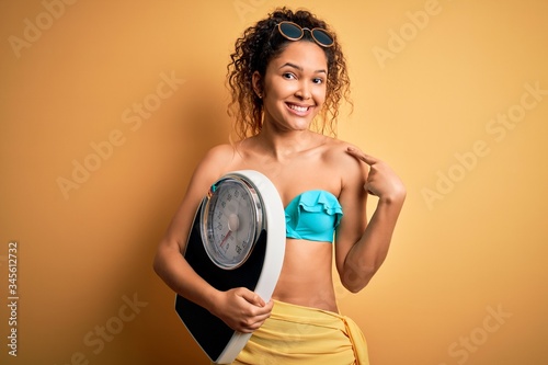 Young beautiful american slim woman on vacation wearing bikini holding weight machine with surprise face pointing finger to himself