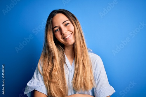 Young beautiful blonde woman with blue eyes wearing striped shirt over blue background happy face smiling with crossed arms looking at the camera. Positive person. © Krakenimages.com