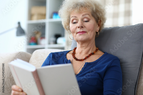 Portrait of well-educated senior woman reading interesting book. Attractive female sitting on comfort couch at home and relaxing. Cozy interior. Hobby and spare time concept photo