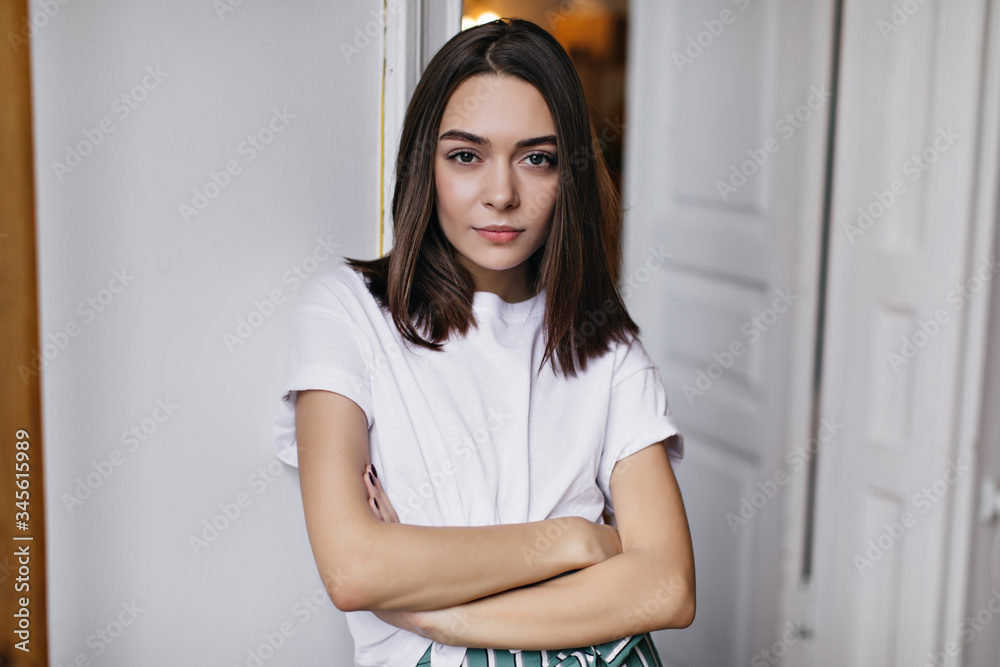 Confident lady with straight hairstyle standing in her apartment with arms crossed. Indoor shot of serious european woman wears white t-shirt. Confident lady with straight hairstyle standing in her apartment with arms crossed. Indoor shot of serious european woman wears white t-shirt.
