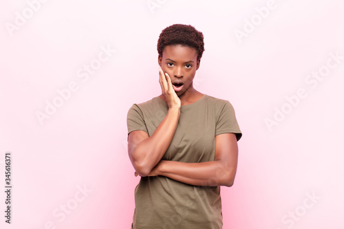 young pretty black womanopen-mouthed in shock and disbelief, with hand on cheek and arm crossed, feeling stupefied and amazed against pink wall photo