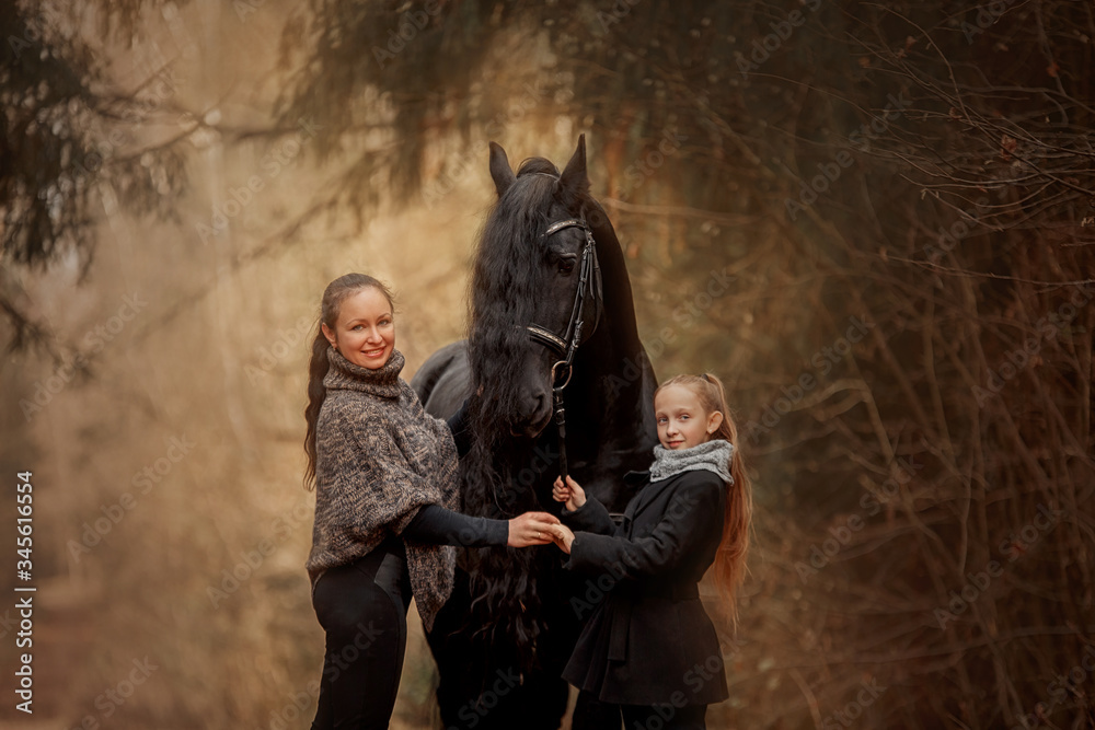 Mother and daughter with Black Beautiful Friesian stallion with long hair outdoor portrait in an autumn forest Mother and daughter with Black Beautiful Friesian stallion with long hair outdoor portrait in an autumn forest