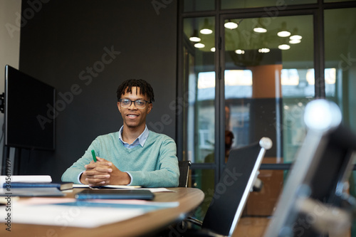 Young African-American man with dreadlocks wearing eyeglasses sitting alone at office board room table looking at camera smiling, copy space