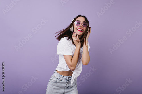 Winsome girl in white t-shirt dancing on purple background and expressing positive emotions. Indoor portrait of pretty dark-haired woman chilling in studio.