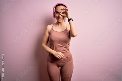 Young fitness woman wearing sport workout clothes wearing headphones listening to music doing ok gesture with hand smiling, eye looking through fingers with happy face.