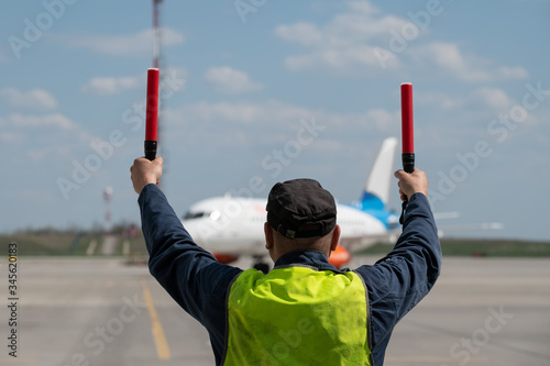 male airport marshal with electric wands in a yellow uniform helps park the plane. The supervisor meets a passenger plane at the airport. The plane steers into the parking lot.