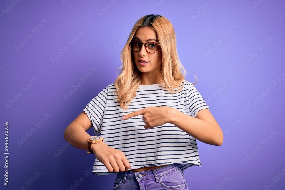 Young beautiful blonde woman wearing striped t-shirt and glasses over purple background In hurry pointing to watch time, impatience, upset and angry for deadline delay Young beautiful blonde woman wearing striped t-shirt and glasses over purple background In hurry pointing to watch time, impatience, upset and angry for deadline delay