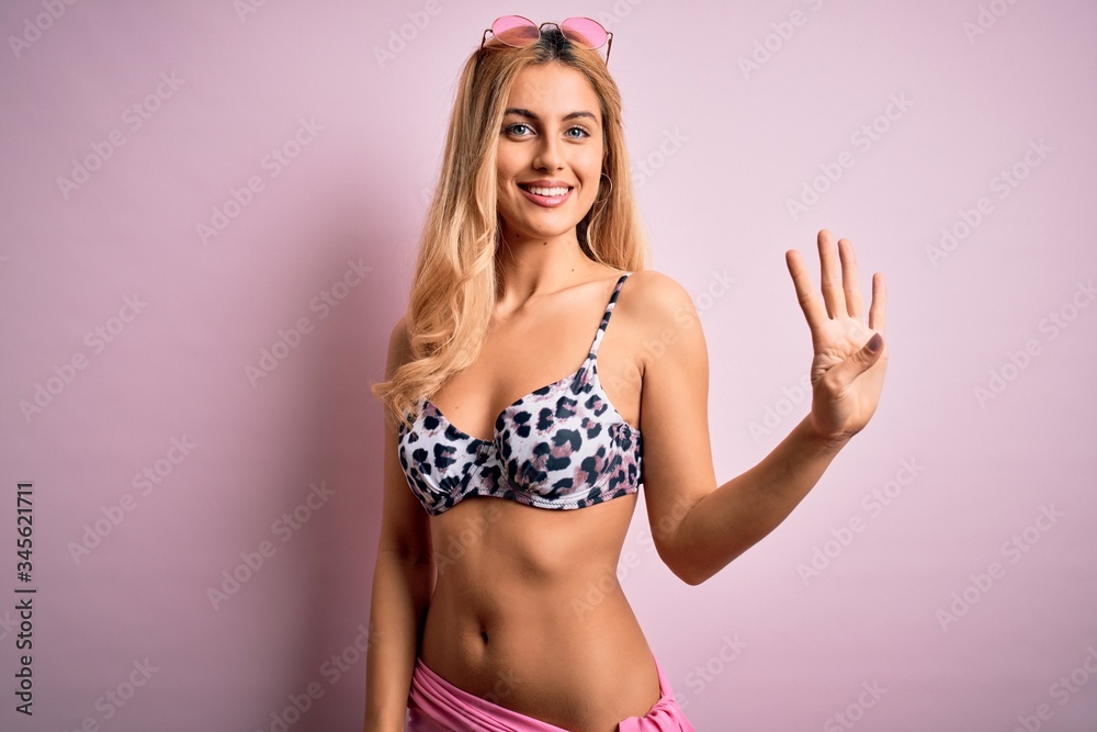 Obraz premium Young beautiful blonde woman on vacation wearing bikini over isolated pink background showing and pointing up with fingers number four while smiling confident and happy.