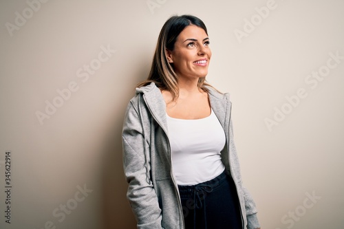 Young beautiful brunette sportswoman wearing sportswoman training over white background looking away to side with smile on face, natural expression. Laughing confident.
