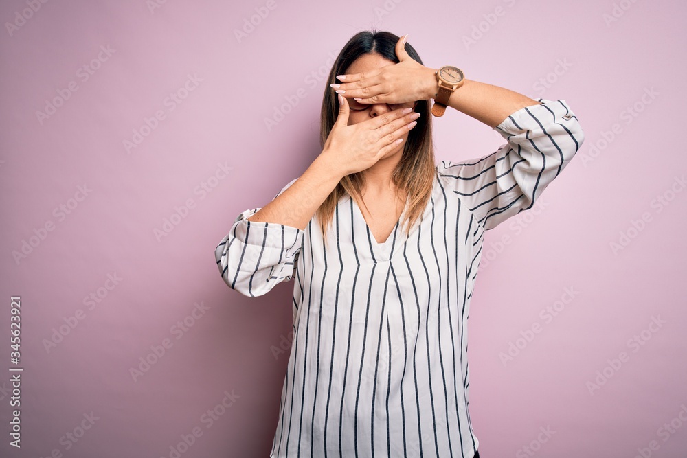 Young beautiful woman wearing casual striped t-shirt and glasses over pink background Covering eyes and mouth with hands, surprised and shocked. Hiding emotion