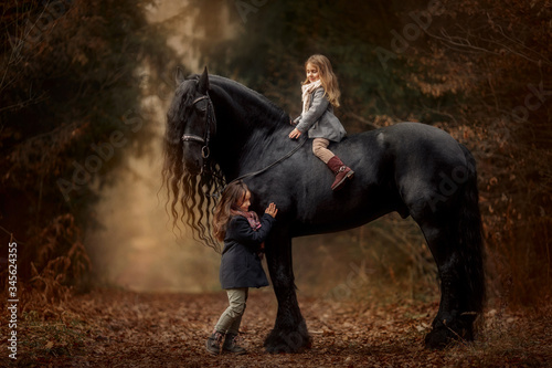 Little sisters with Friesian stallion with long hair outdoor portrait in an autumn forest