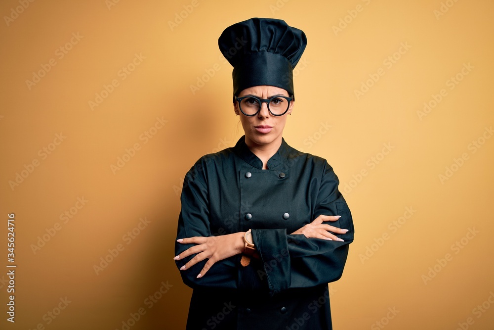 Young beautiful brunette chef woman wearing cooker uniform and hat over yellow background skeptic and nervous, disapproving expression on face with crossed arms. Negative person. Young beautiful brunette chef woman wearing cooker uniform and hat over yellow background skeptic and nervous, disapproving expression on face with crossed arms. Negative person.