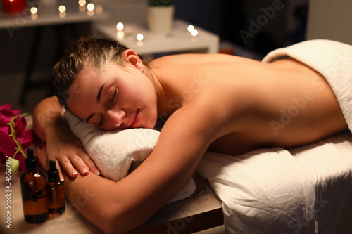 Close-up of glad smiling young woman covered with white towel in spa salon. Bottles with aromatic oils on table. Beauty day. Wellness and massage cabinet concept