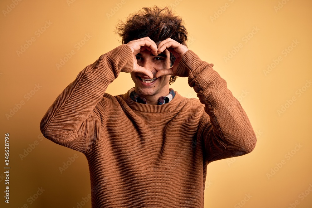 Young handsome man wearing casual shirt and sweater over isolated yellow background Doing heart shape with hand and fingers smiling looking through sign Young handsome man wearing casual shirt and sweater over isolated yellow background Doing heart shape with hand and fingers smiling looking through sign