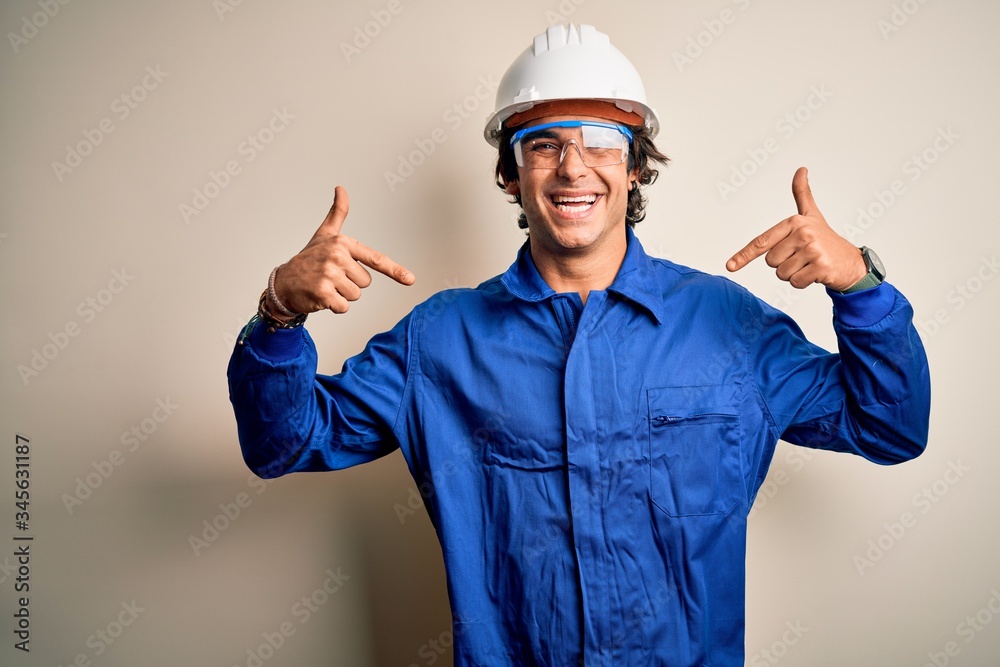 Young constructor man wearing uniform and security helmet over isolated white background looking confident with smile on face, pointing oneself with fingers proud and happy.
