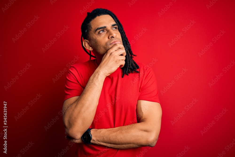 Young handsome african american afro man with dreadlocks wearing red casual t-shirt with hand on chin thinking about question, pensive expression. Smiling with thoughtful face. Doubt concept.