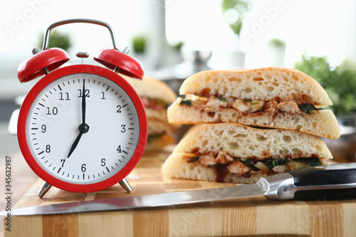 Close-up of red clock showing seven in morning. Hot sandwich with tasty stuffing on table. Quick breakfast before work. Unhealthy eating. Hurry up and fast food concept