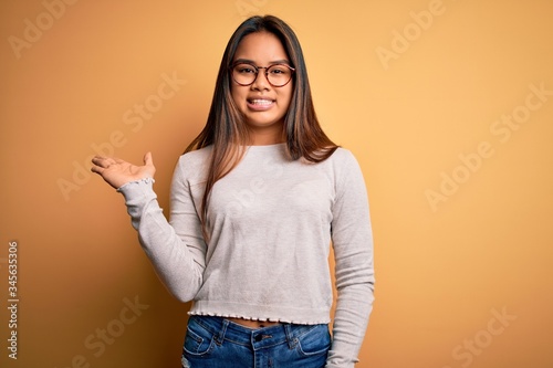 Young beautiful asian girl wearing casual sweater and glasses over yellow background smiling cheerful presenting and pointing with palm of hand looking at the camera.
