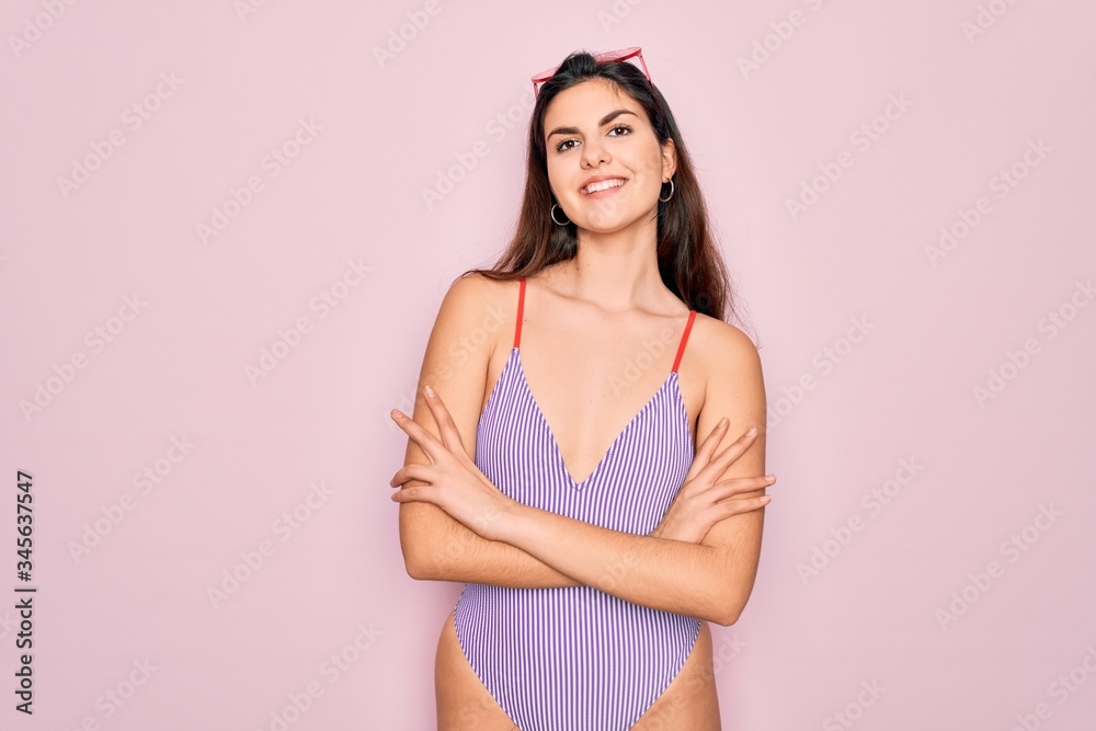 Young beautiful fashion girl wearing swimwear swimsuit and sunglasses over pink background happy face smiling with crossed arms looking at the camera. Positive person. Young beautiful fashion girl wearing swimwear swimsuit and sunglasses over pink background happy face smiling with crossed arms looking at the camera. Positive person.