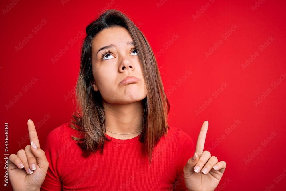 Young beautiful brunette girl wearing casual t-shirt over isolated red background Pointing up looking sad and upset, indicating direction with fingers, unhappy and depressed.