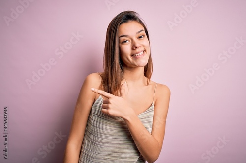 Young beautiful brunette girl wearing casual striped t-shirt over isolated pink background cheerful with a smile of face pointing with hand and finger up to the side with happy and natural expression