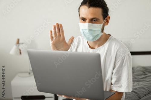 Man in medical mask is working from home with a laptop and speaking to his friends or business partners via webcam because of coronavirus.