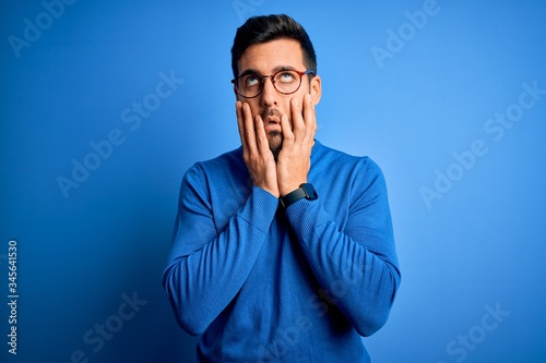 Young handsome man with beard wearing casual sweater and glasses over blue background Tired hands covering face, depression and sadness, upset and irritated for problem