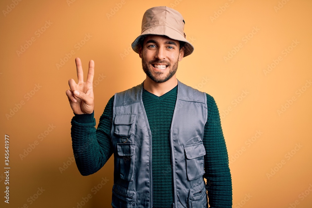 Handsome tourist man with beard on vacation wearing explorer hat over yellow background showing and pointing up with fingers number two while smiling confident and happy.